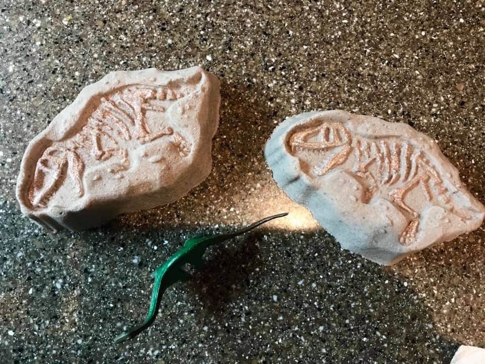 Two pale dino fossil casts on grit beside a small green dinosaur toy