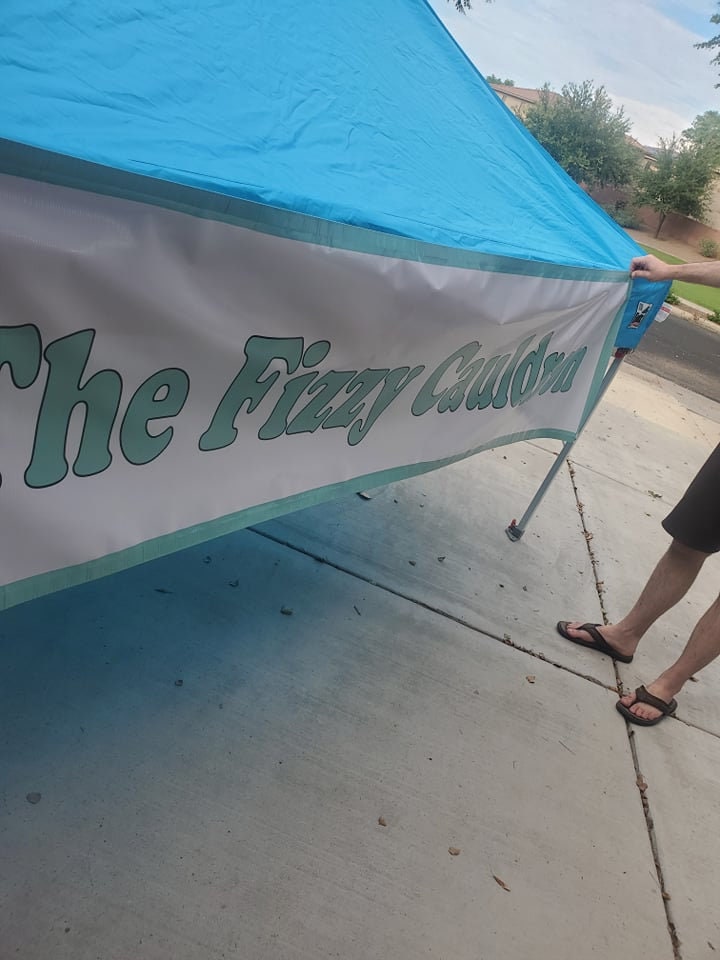 The Fizzy Cauldron banner tied along the edge of a blue market canopy at an outdoor stall.