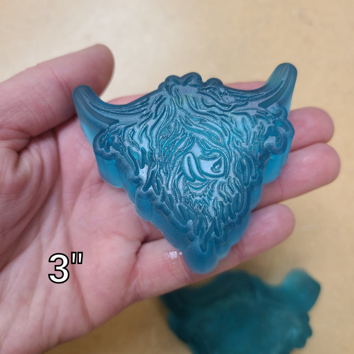 Translucent teal highland cow head cast held in a palm and marked three inches.