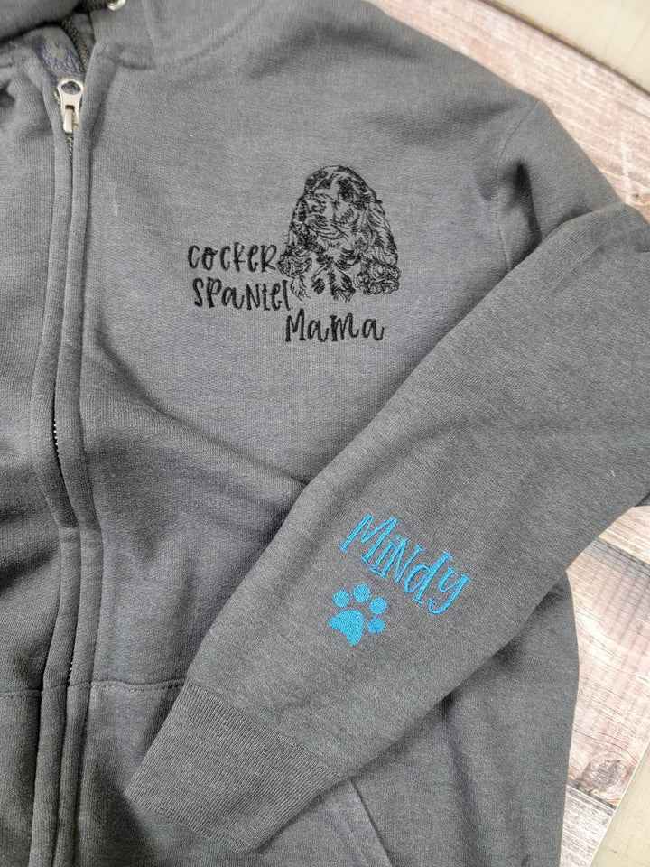 Folded grey zip hoodie showing the cocker spaniel chest embroidery and a blue name on the sleeve.