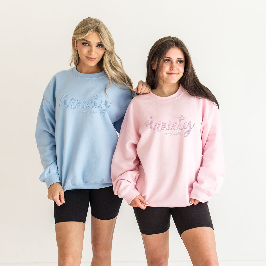 Two models in light blue and pink crewnecks, each embroidered with Anxiety in tonal script.