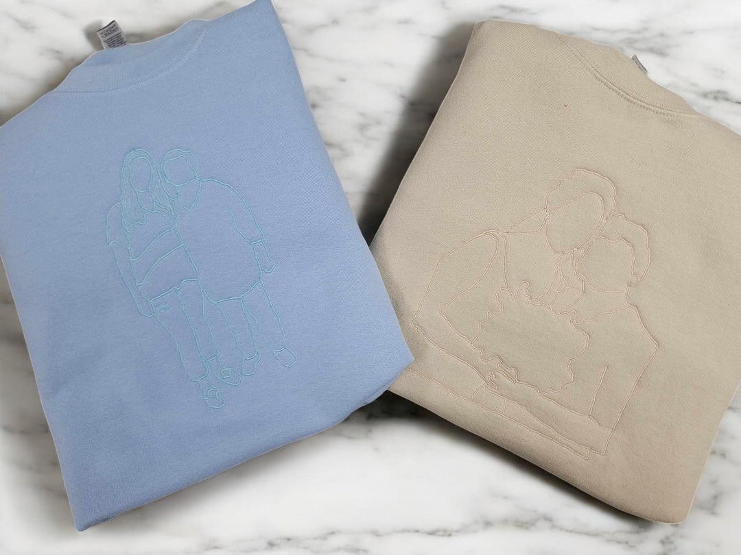Light blue and sand crewnecks folded on marble, each embroidered with a tone-on-tone photo outline.