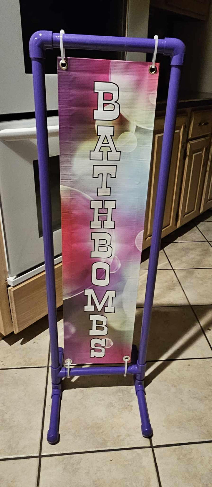 Vertical BATHBOMBS banner hung inside a purple PVC display frame in a kitchen.