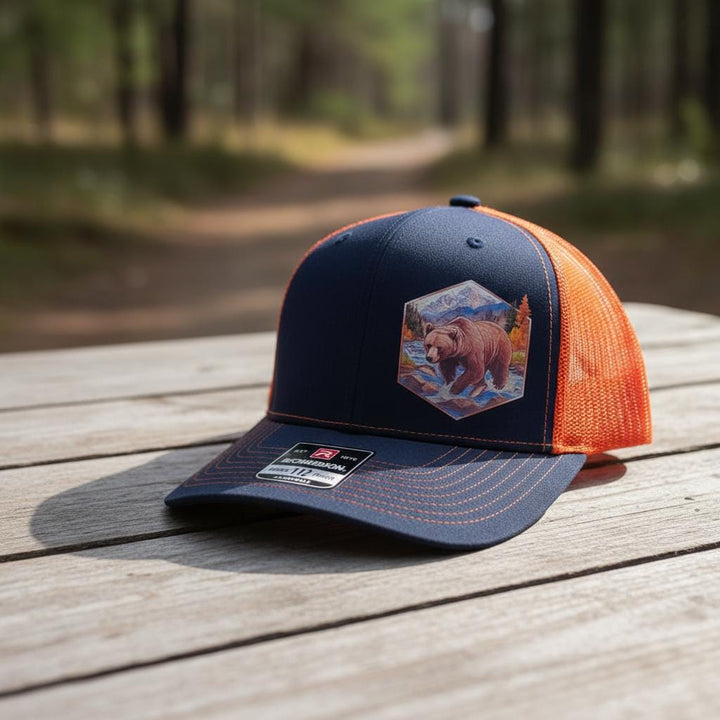 Navy and orange trucker cap with a hexagon bear patch, on a picnic table in the woods.