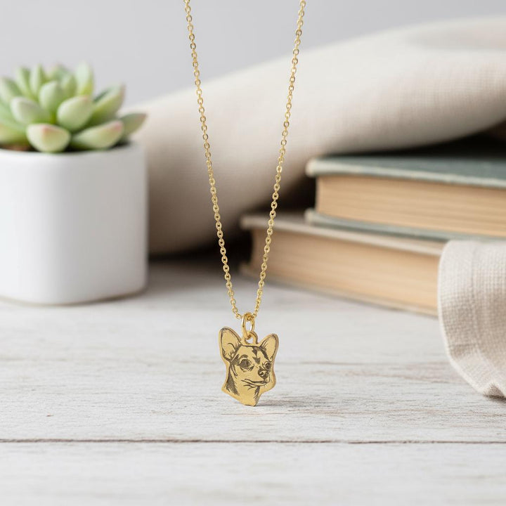 Gold chain laid flat on white boards with the Chihuahua pendant centred, books behind.