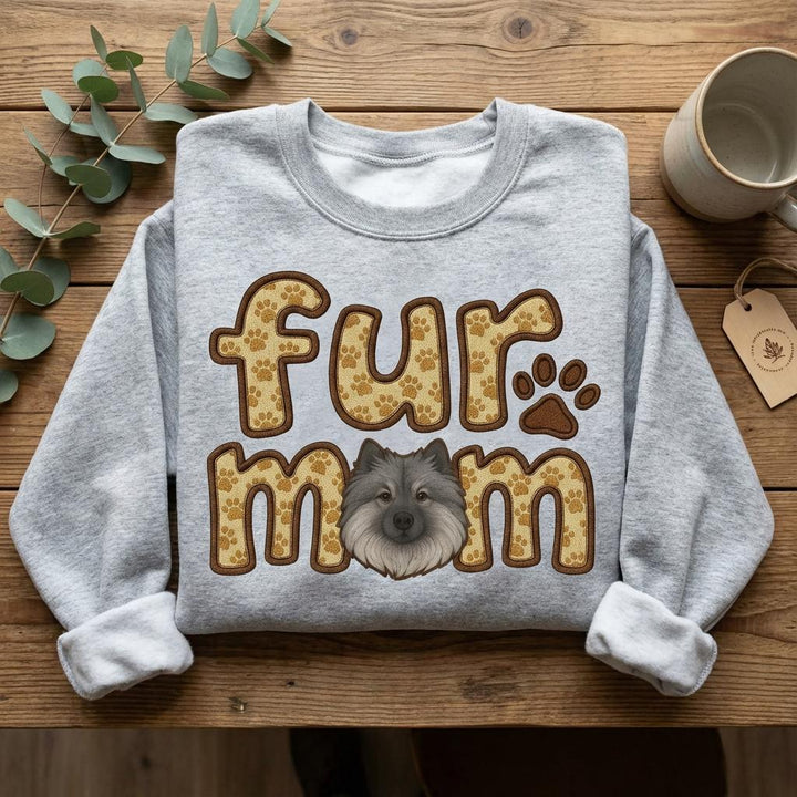 Grey sweatshirt flat on wood with the fur mom Keeshond print, eucalyptus and a mug alongside.