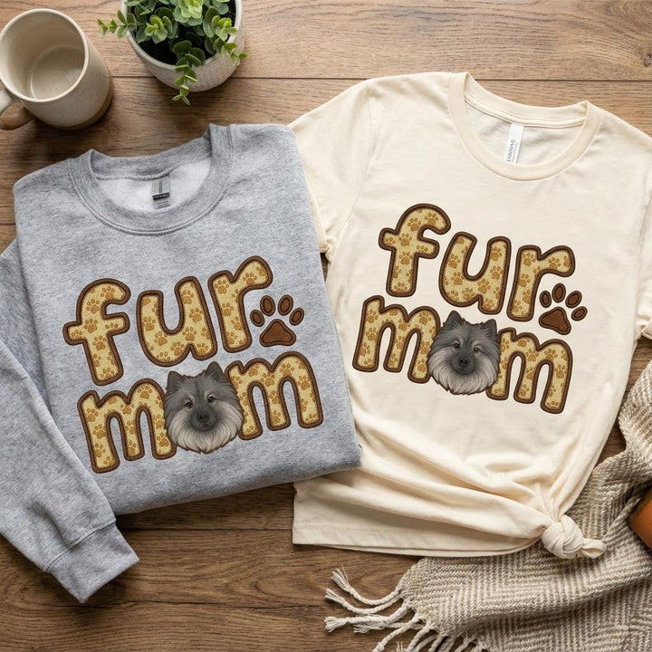 Grey sweatshirt and cream tee side by side on wood, both carrying the fur mom Keeshond print.