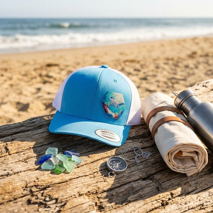 Blue manta ray trucker hat on driftwood with sea glass, a compass and a rolled towel.