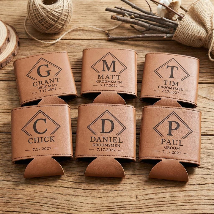 Six engraved leatherette can coolers in a flat lay on wood with twine and dried stems.