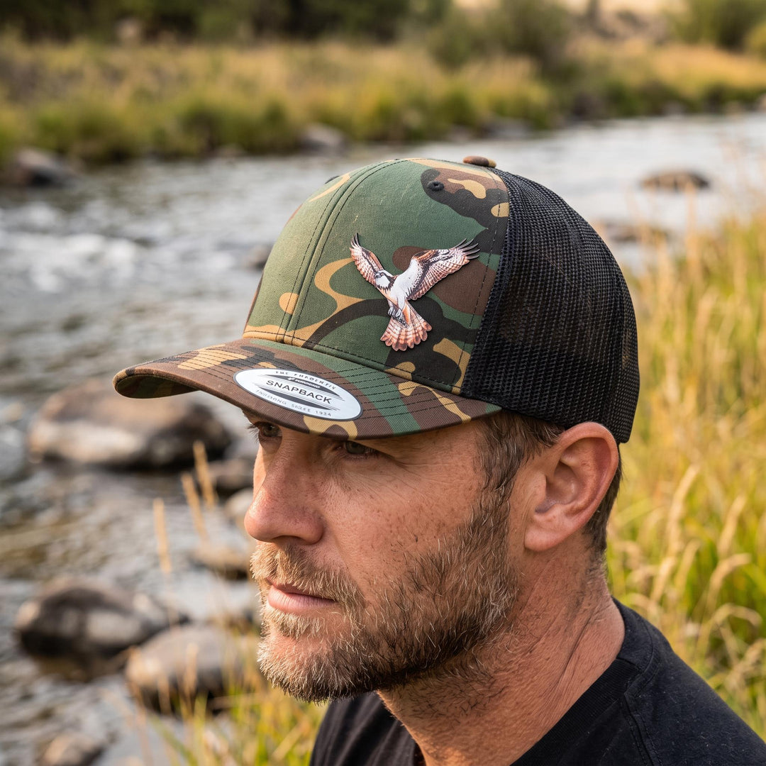 Man by a river wearing the woodland camo trucker hat with the osprey patch.