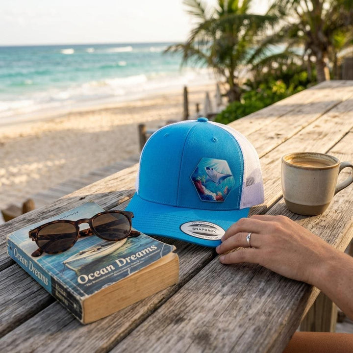 Blue manta ray trucker hat on a beachside table with sunglasses, a mug and a book.