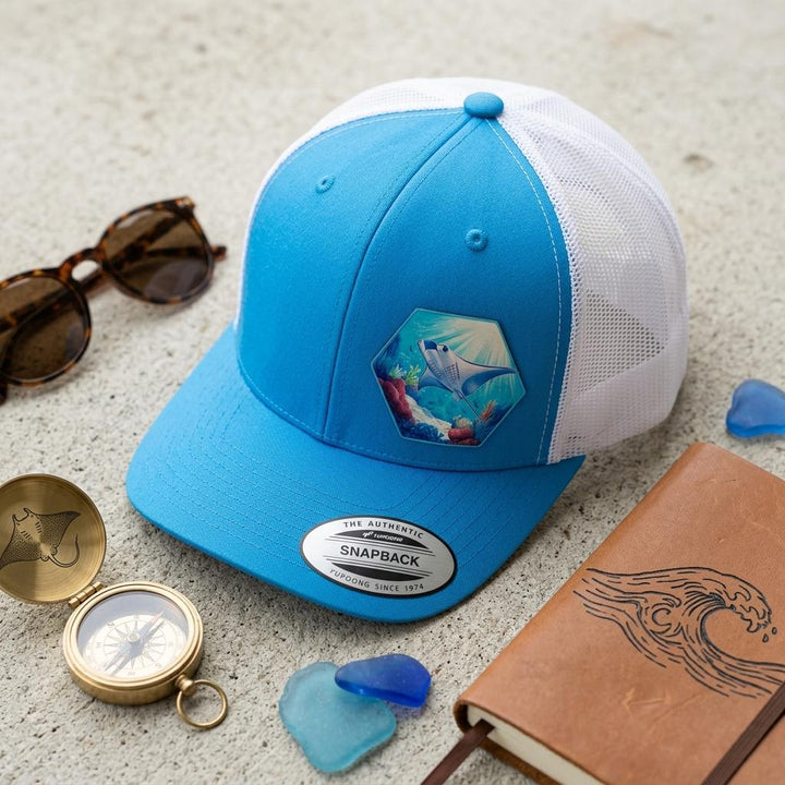 Manta ray reef patch cap on sand beside sunglasses, a compass and a leather notebook.